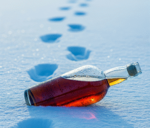 A tipped brandy bottle lies in icy blue snow, with footprints leading away into the distance.