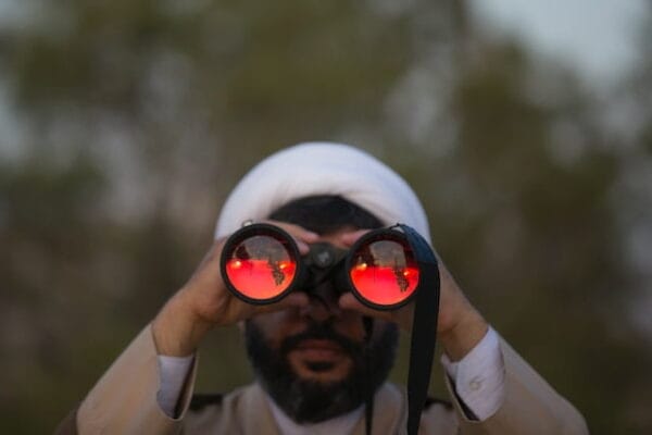 A man wearing a white turban type hat looking through high quality binoculars.Photo by mostafa meraji on Unsplash
