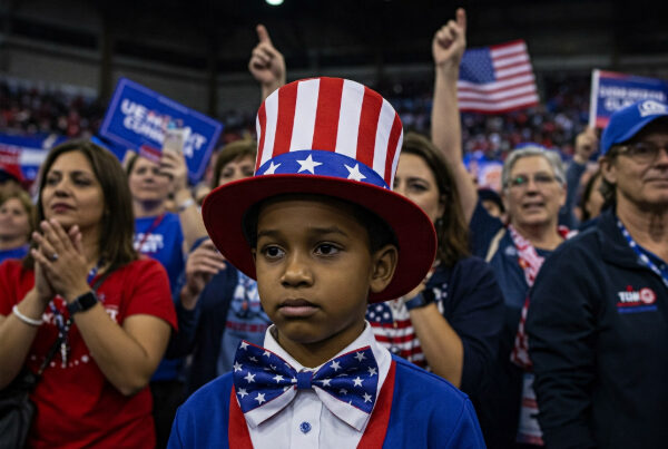 A young, brown-skinned boy at a Democratic rally looks sad, wearing a stars-and-stripes top hat and bow tie. The background is a crowded rally