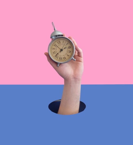 The hand of a white male potruding through a hole on a white background as he holds up an alarm clock an a pink background