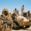 Soldiers in desert camouflage with armored vehicles and sniper under blue sky.
