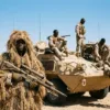 Soldiers in desert camouflage with armored vehicles and sniper under blue sky.