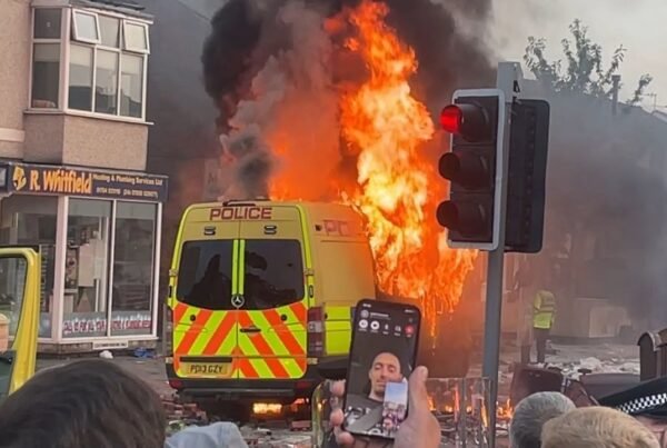 A police van on fire while a man takes a selfie in Southport 2024