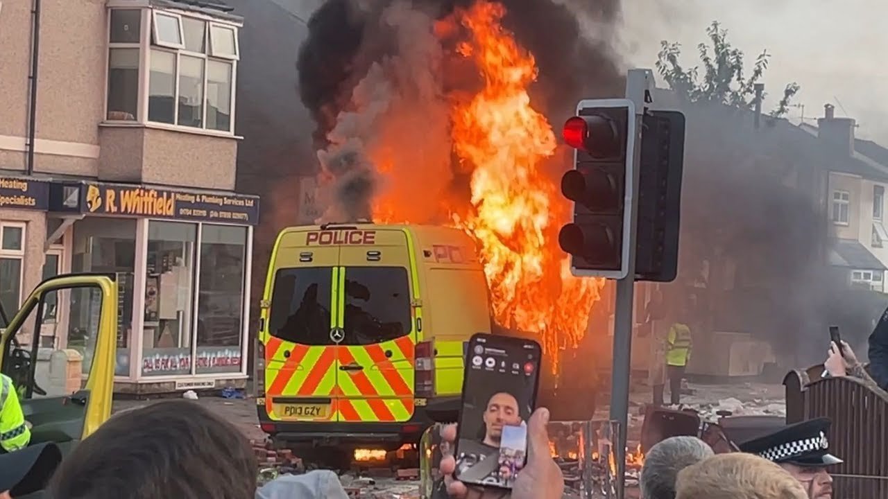 A police van on fire while a man takes a selfie in Southport 2024