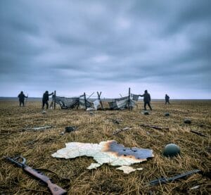 A mowed, weapon-strewn field with a burnt Ukraine map. Shadowy figures fail at a barrier under a grim, overcast sky.