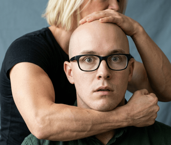 Close-up of a shocked young bald man with glasses, held tightly around the neck from behind by a muscular arm.